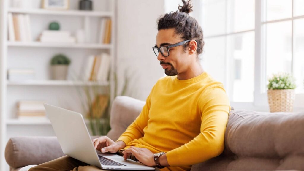 Create product listings A man in a yellow sweater sitting on a couch and working on a laptop.