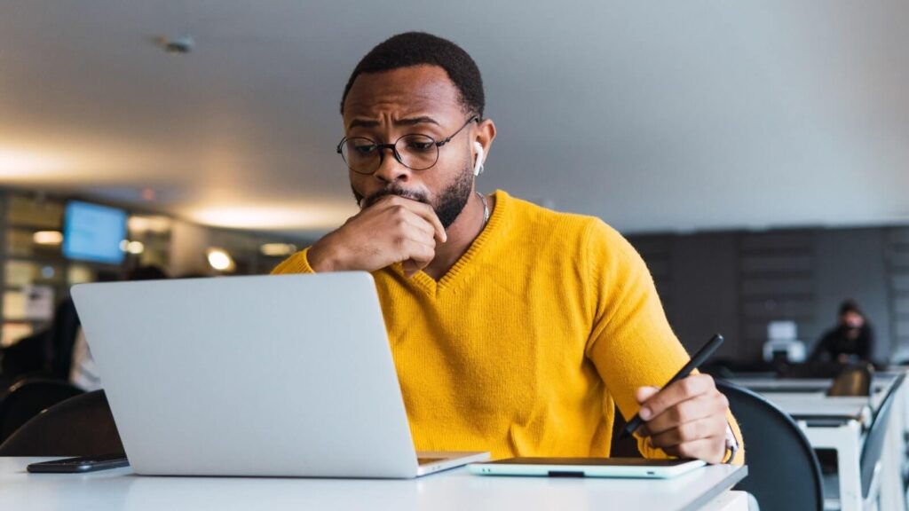 How to sell on Amazon Conduct market research A man in glasses wearing a yellow sweater, looking at a laptop screen and thinking.