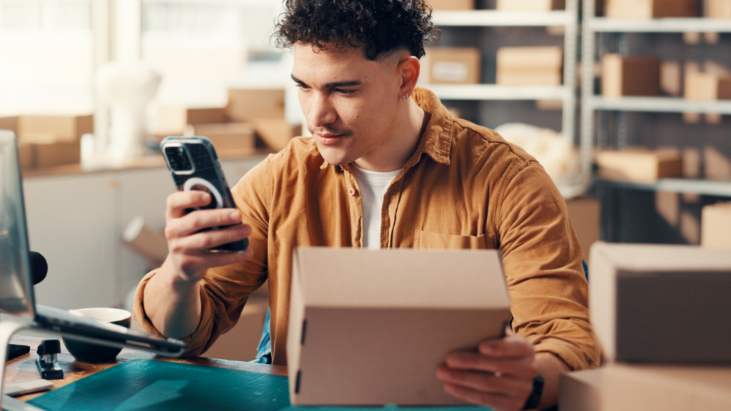 Choose your fulfillment model and supplier A man in a brown shirt is in a workspace, holding a smartphone and looking at a cardboard box.