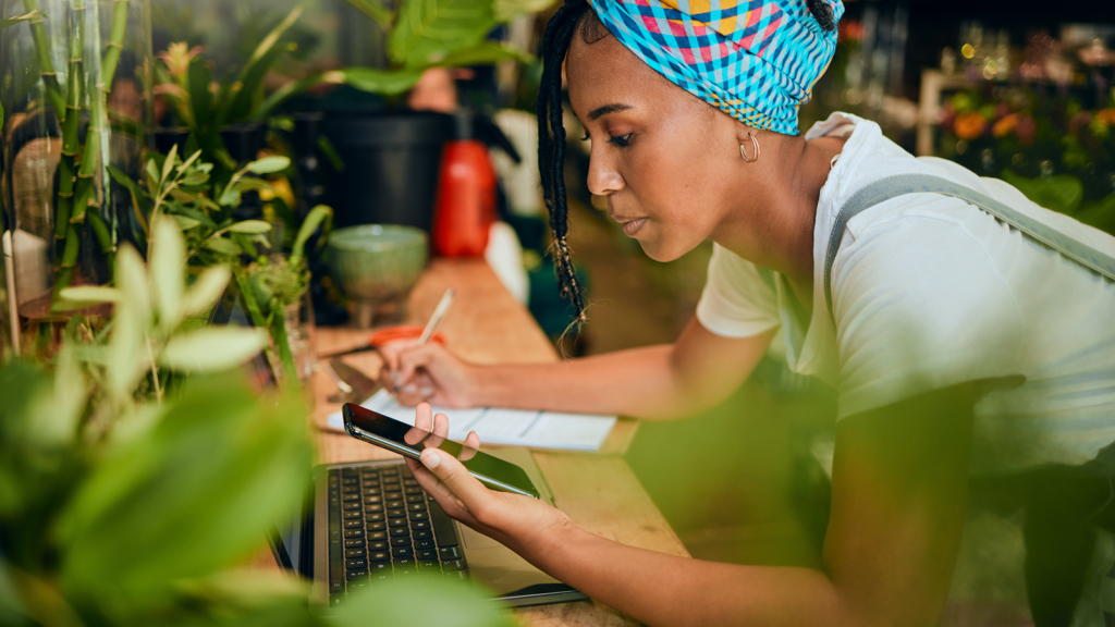 Common challenges and risks in TikTok dropshipping A woman in a colorful headscarf leans over plants, using a smartphone and laptop, taking notes.