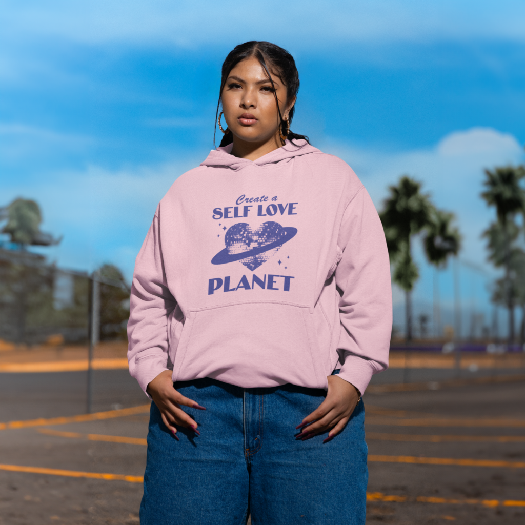 Positive affirmation messages A woman in a pink “Create a self love planet” custom hoodie and blue jeans standing in a parking lot.