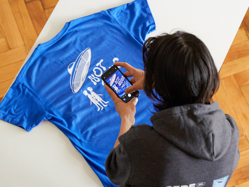 Sell on TikTok with Printify A person in a gray hoodie photographs a blue t-shirt featuring an alien and UFO design with "NOT ALONE" text.