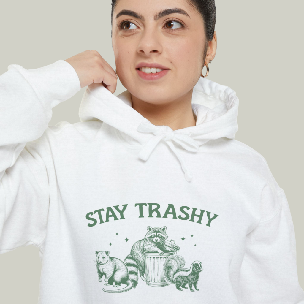Unisex garment dyed hoodie Comfort Colors 1567 A close-up shot of a woman wearing a white “Stay Trashy” hoodie with an illustrated raccoon and other animals.