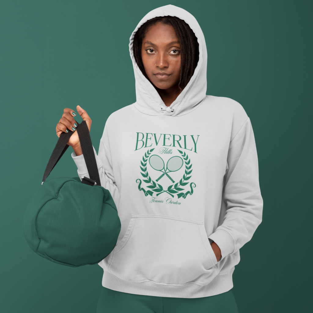 Vintage sports logos A woman in a white "Beverly Hills Tennis Club" hoodie holding a green bag against a green background.